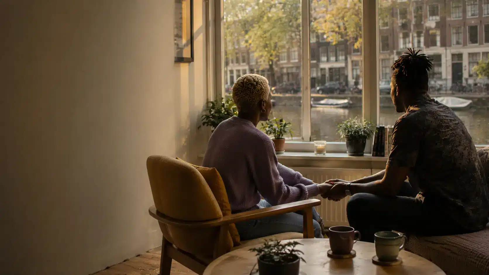Two people hold hands while talking in a bright room overlooking an Amsterdam canal.